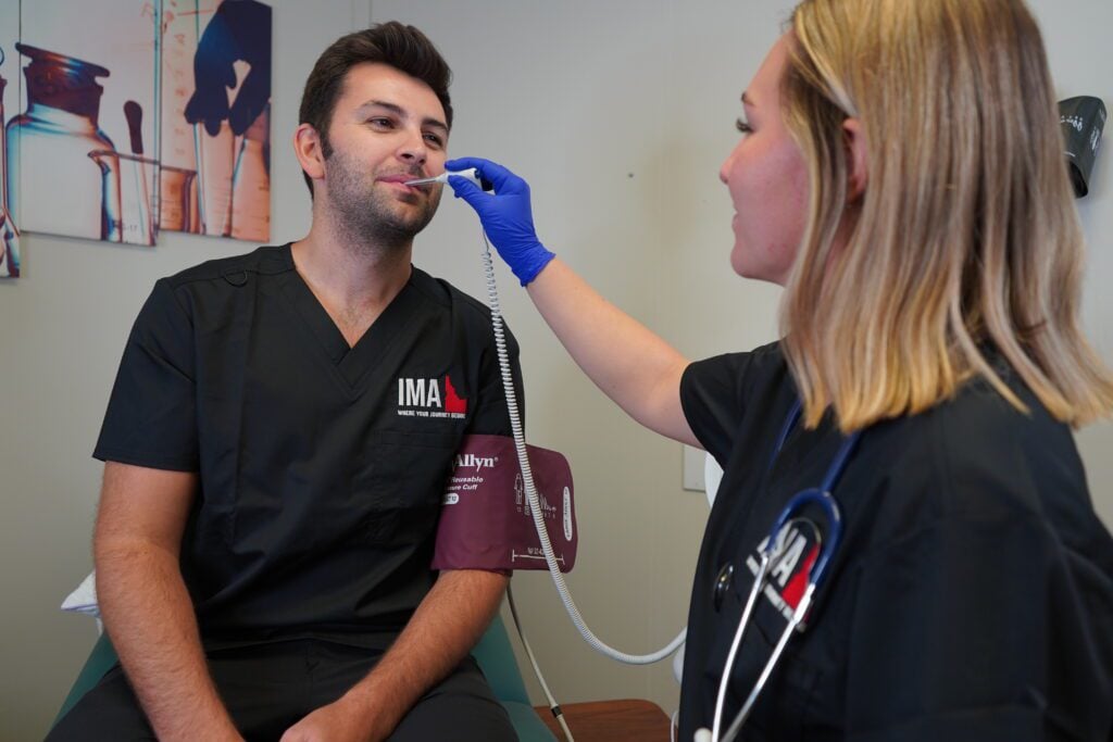 An IMA Medical Assistant course student takes another student's temperature in a clinical setting to practice hands-on skills.