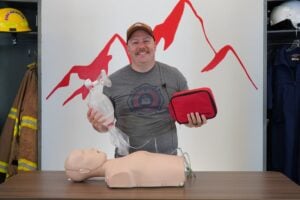 EMT instructor holding EMT airway management equipmment, standing in front of IMA mountain logo with fire gear and flight EMS gear flanking him on the sides.