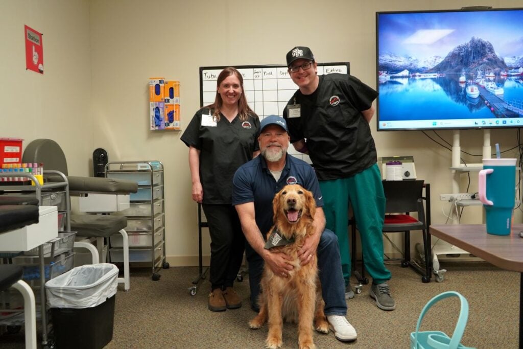 Instructors and students from Idaho Medical Academy's Phlebotomy Technician Program stand in a classroom with a golden retriever sitting politely in front of them.